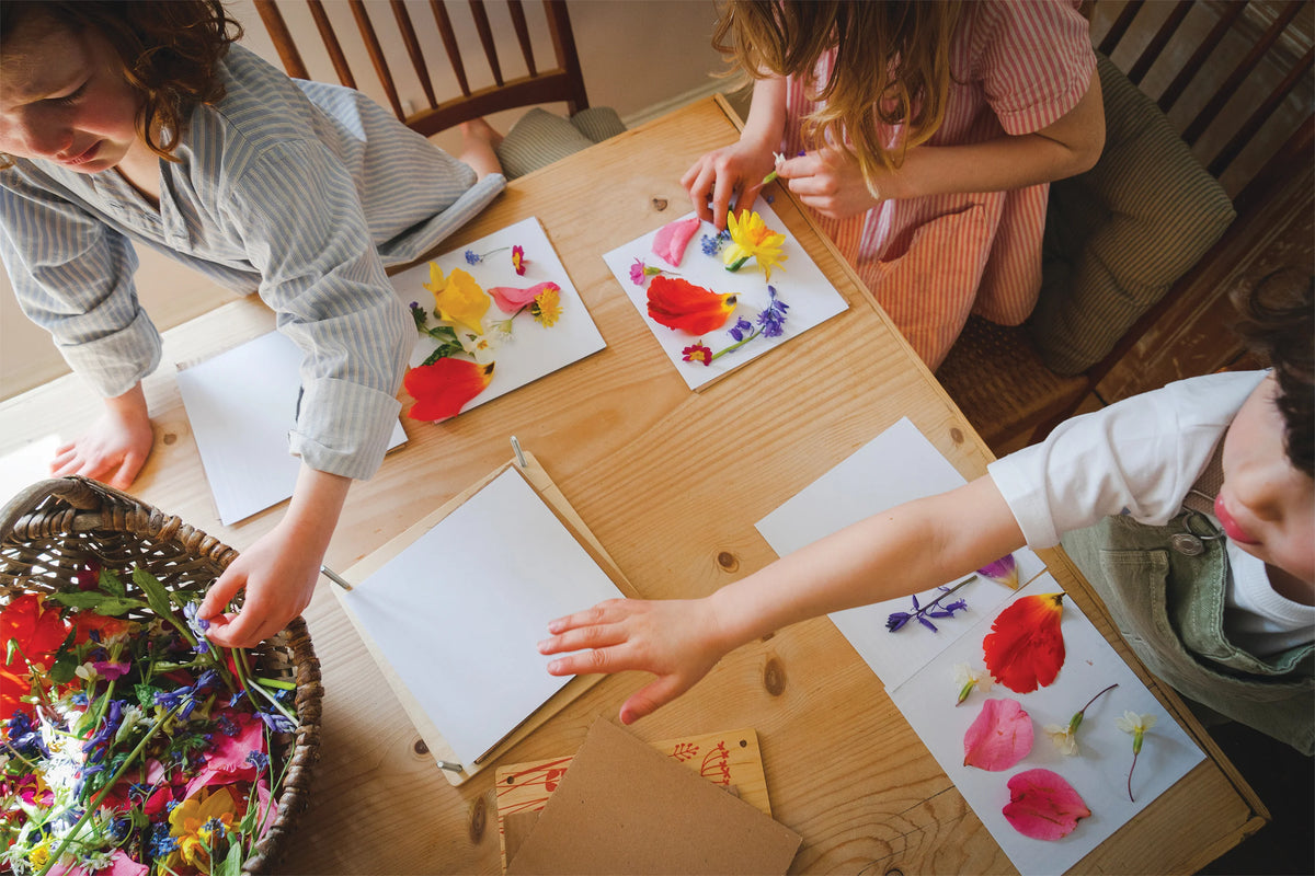 Three children at a table pressing gathered flowers onto paper sheets with the botanical press