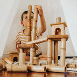 Q Toys Bamboo Building Set, child stacking bamboo lengths into a tower with small wooden houses · view 1