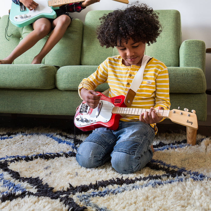 A child sitting on a rug playing a small red and white electric guitar