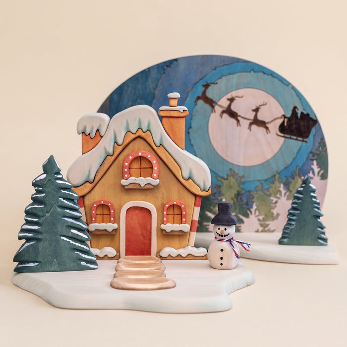 Wooden Christmas scene with a snow-covered gabled house, a moon panel behind it, and small snow trees