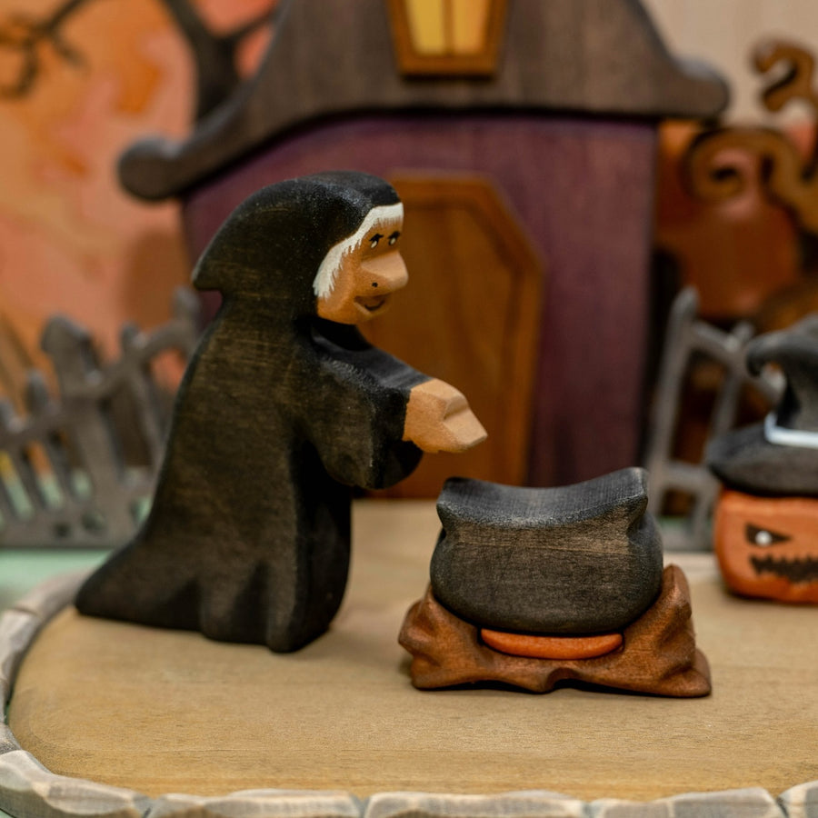Wooden hooded witch leaning over a black cauldron that sits on a carved wooden fire base