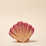 Wooden scallop shell painted pink, standing upright on sand · view 1