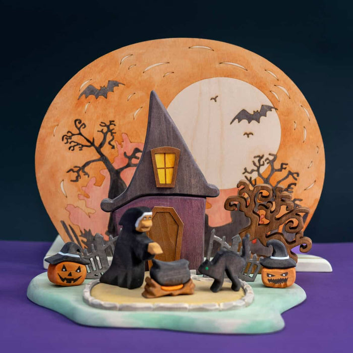 Wooden Halloween scene with a witch house, a full moon panel behind it, a fence, and small pumpkins